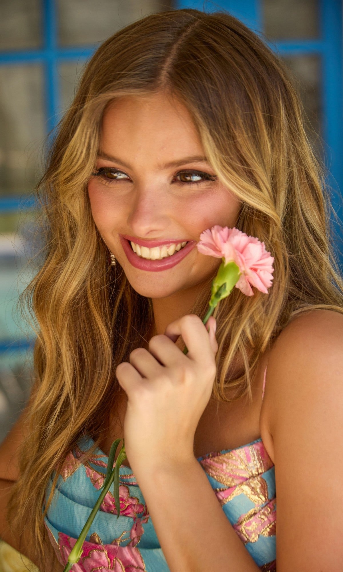 A woman with long wavy hair smiles while holding a pink flower near her face. She is wearing the Ava Presley 42462 Short Homecoming Dress, featuring a colorful, fit-and-flare silhouette with a floral jacquard pattern.